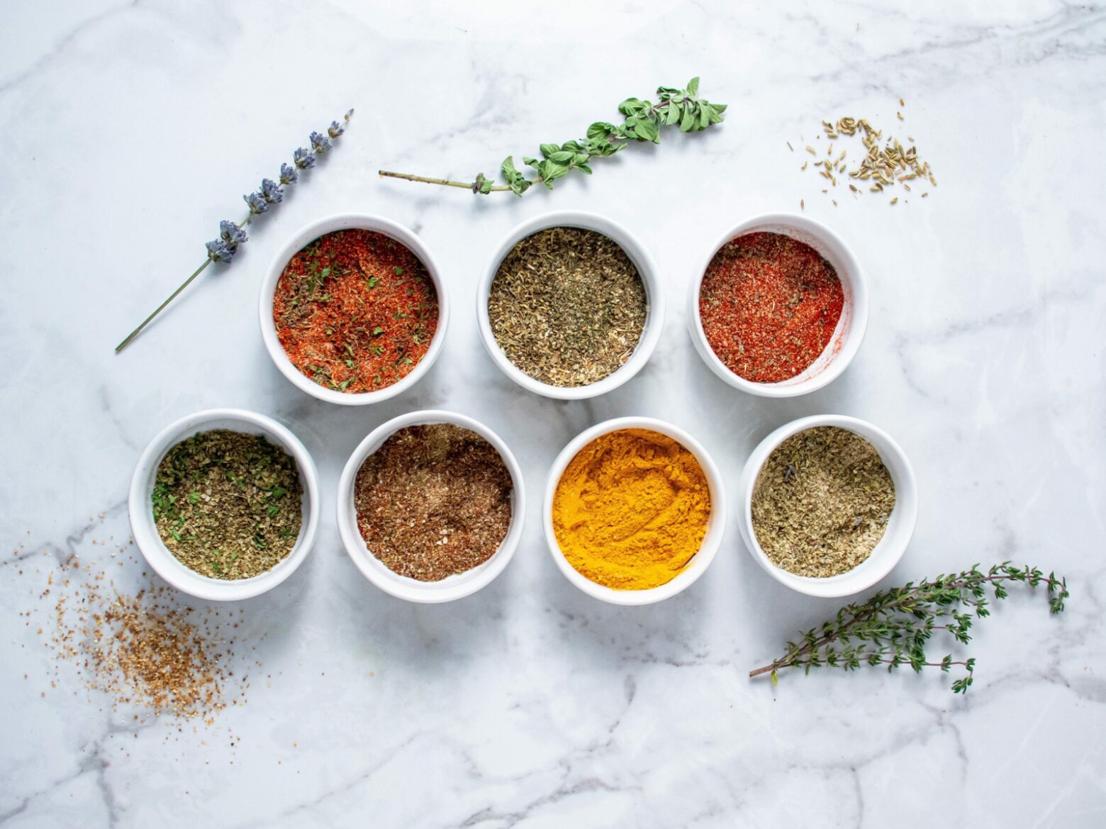 From above, seven small white bowls containing different spice blends with herbs and seeds surrounding them.