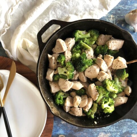 An iron skillet containing stir-fried chicken and broccoli next to an empty white plate with a knife and fork and a bulb of garlic.