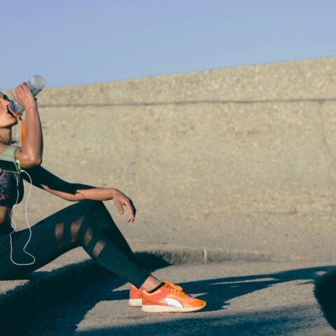 A woman in running shoes and related running gear drinks water on a step.