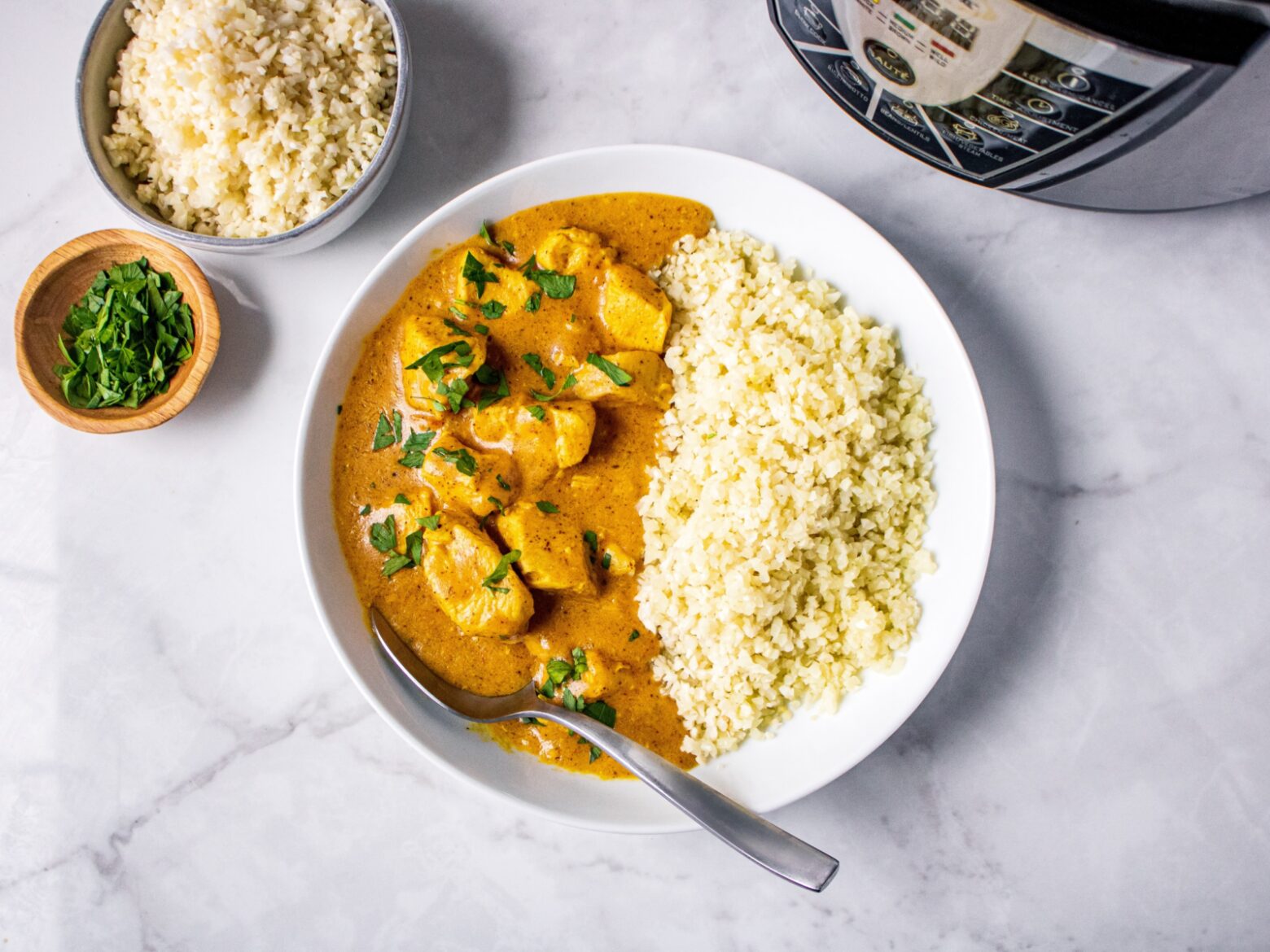 From above, a bowl of half butter chicken garnished with chopped parsley with a silver spoon and half cauliflower rice.