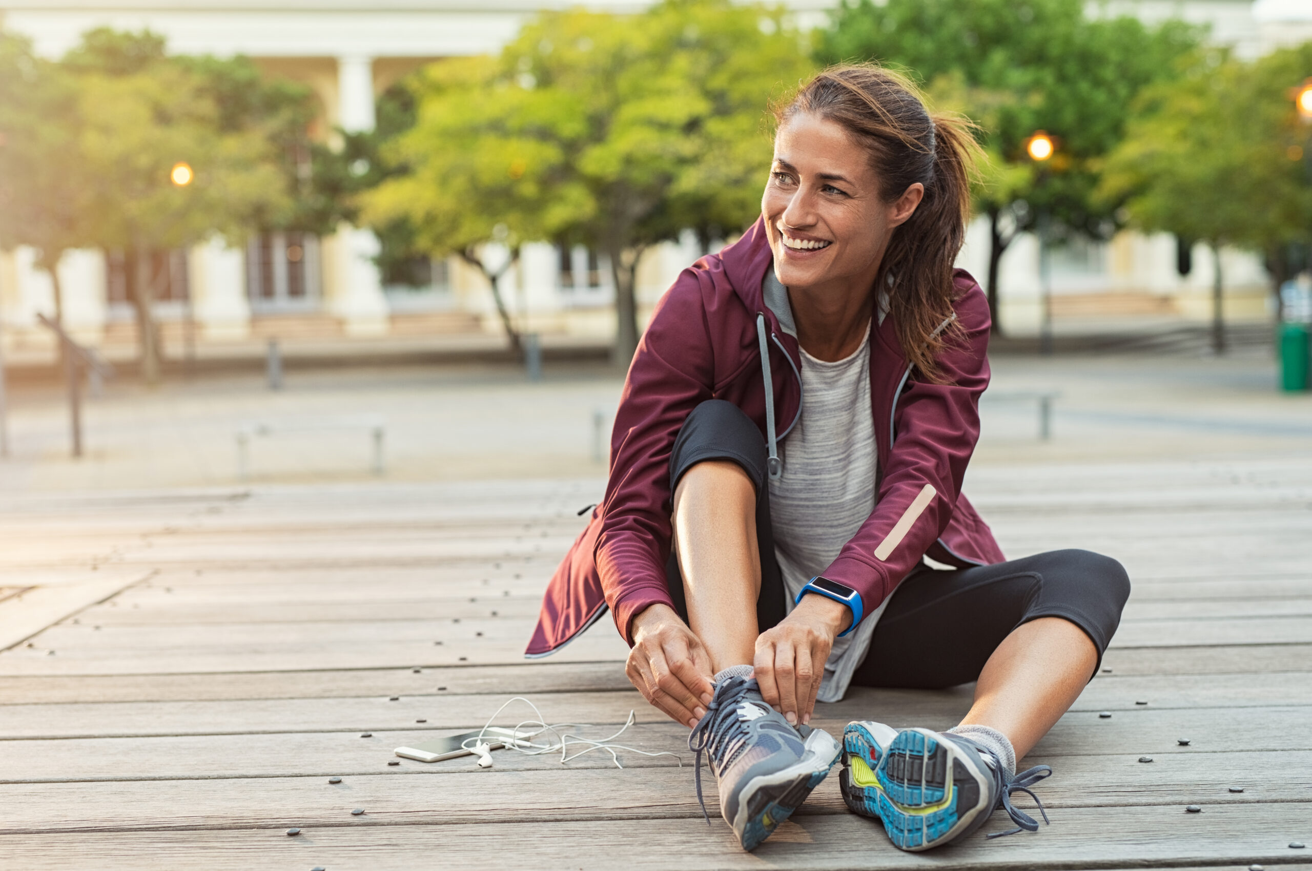 Sporty woman in maroon hoodie smiles and adjusts her shoes as she prepares for a run.