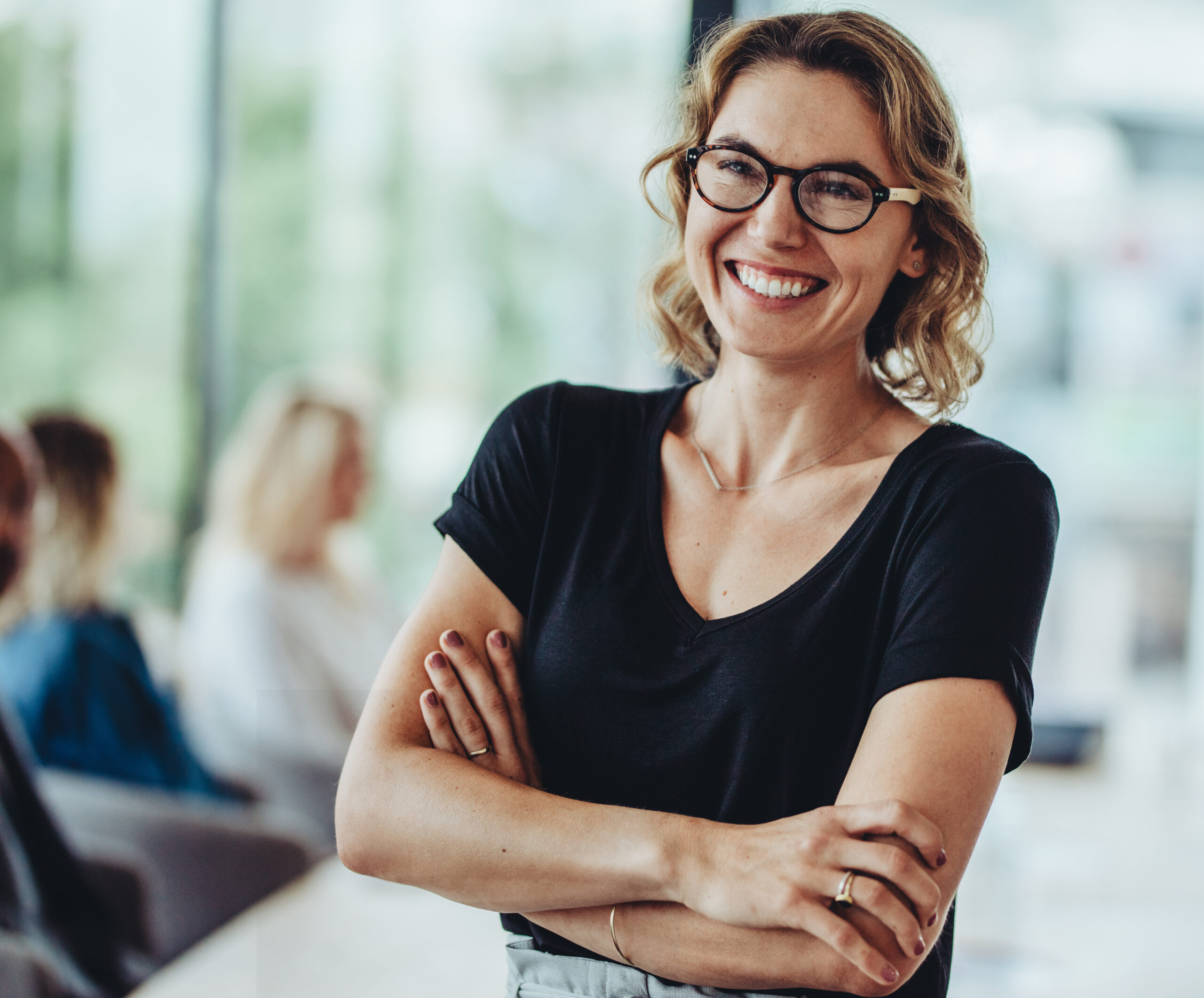 Portrait of smiling businesswoman standing in office with colleagues meeting in background. Successful female professional with her arms crossed in meeting room.