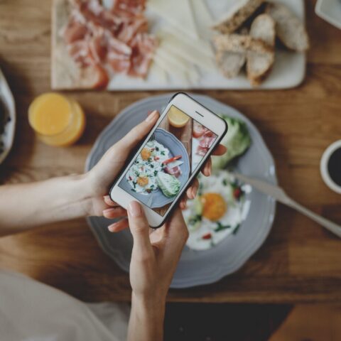 From above, a woman photographs her meal on a table surrounded by food.