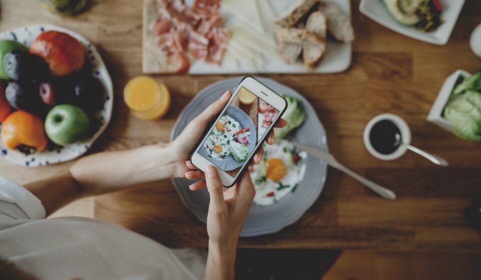 From above, a woman photographs her meal on a table surrounded by food.